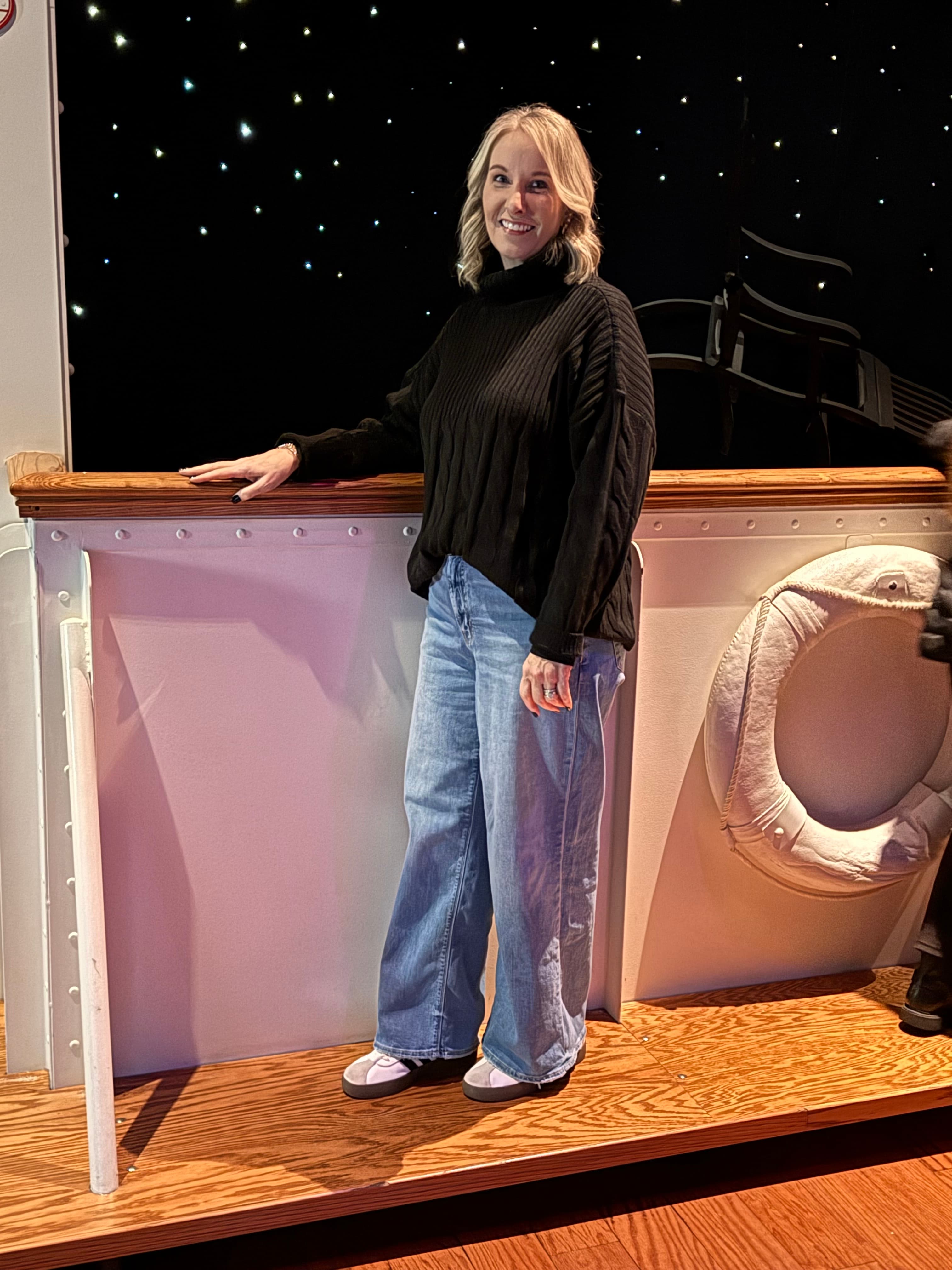 Smiling blonde woman in a black sweater and jeans on a starry ship deck.