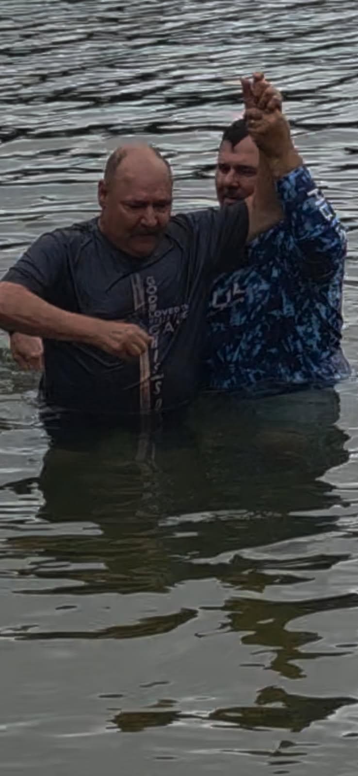 Two men waist-deep in water during a baptism, one holding the other's hand high.